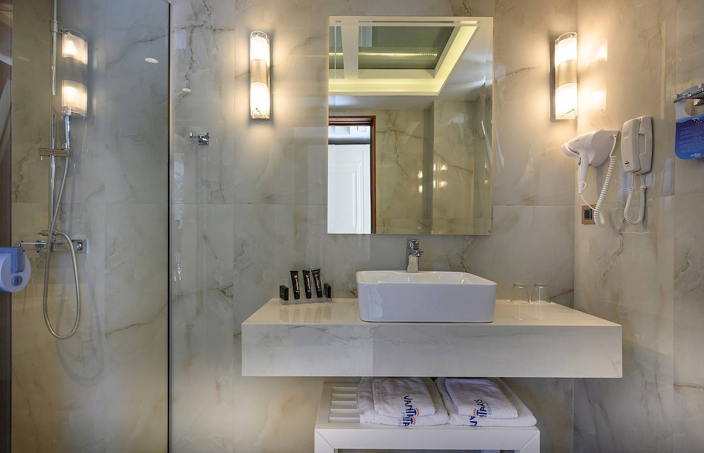 Badezimmer im Vantaris Palace Hotel mit Marmorfliesen, Waschbecken, Spiegel und Föhn.