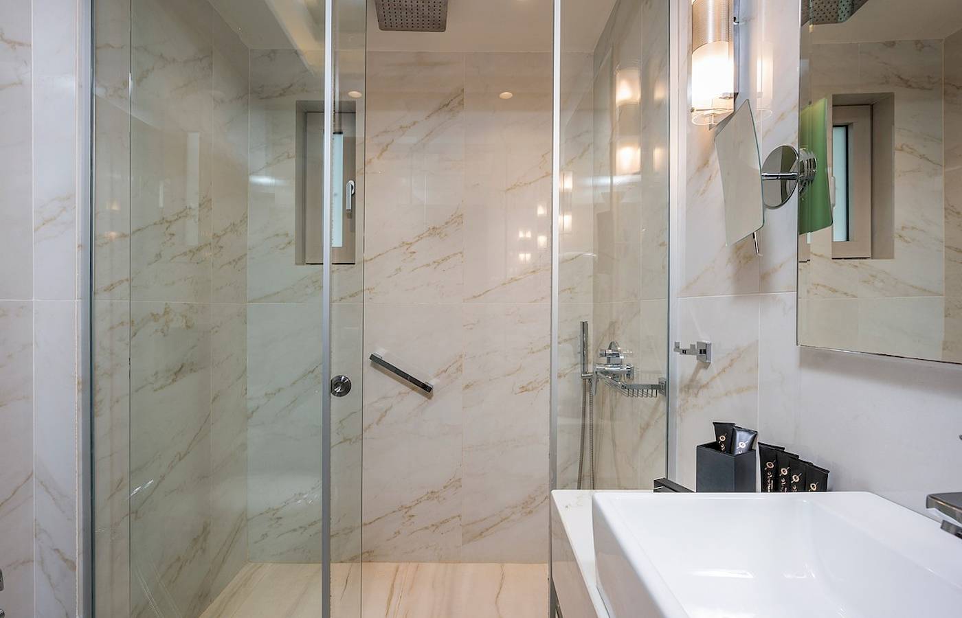 Badezimmer im Vantaris Palace Hotel mit Marmorwänden, Glasdusche und Waschbecken.