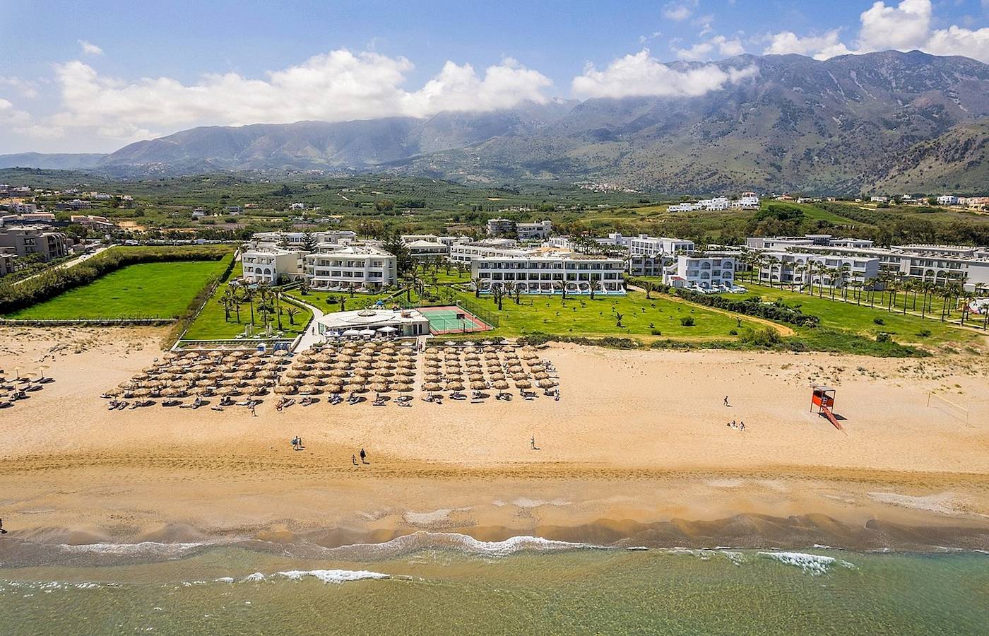 Luftaufnahme des Vantaris Palace Resorts am Strand mit vielen Sonnenschirmen und Bergen im Hintergrund.