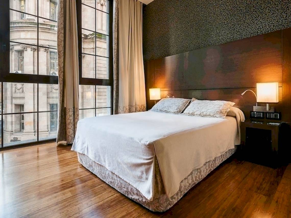 Schlafzimmer im Colonial Barcelona Hotel mit großem Bett, Nachttischen und Fenstern mit Blick auf ein Gebäude.