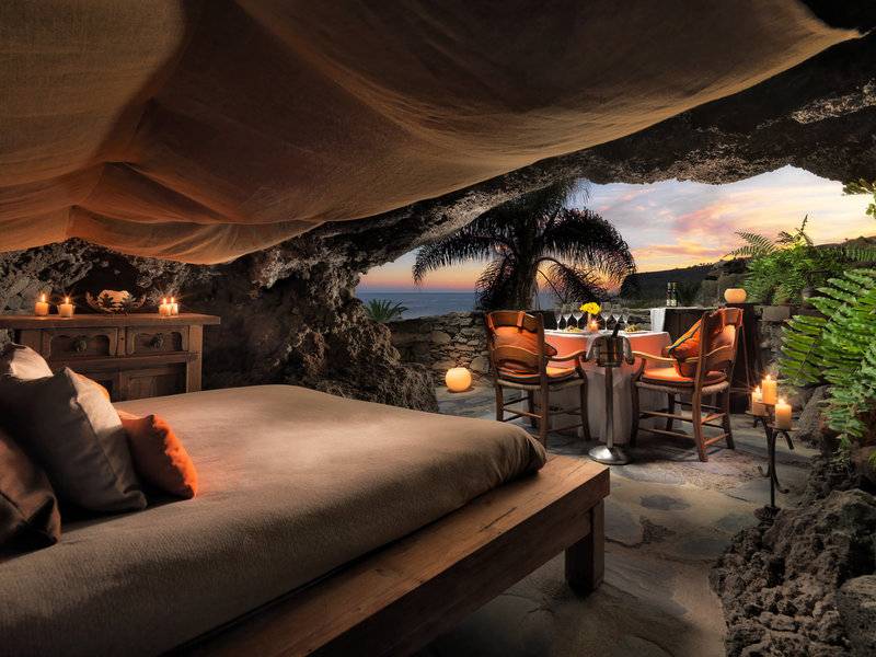 Ein Schlafzimmer, das in einen Felsen gehauen ist, mit Blick auf den Ozean und einem Esstisch mit Kerzen davor.