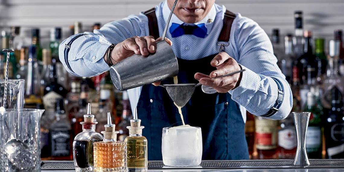 Ein Barkeeper des Myconian Ambassador Resorts bereitet einen Cocktail mit einem Sieb in einer Bar zu.