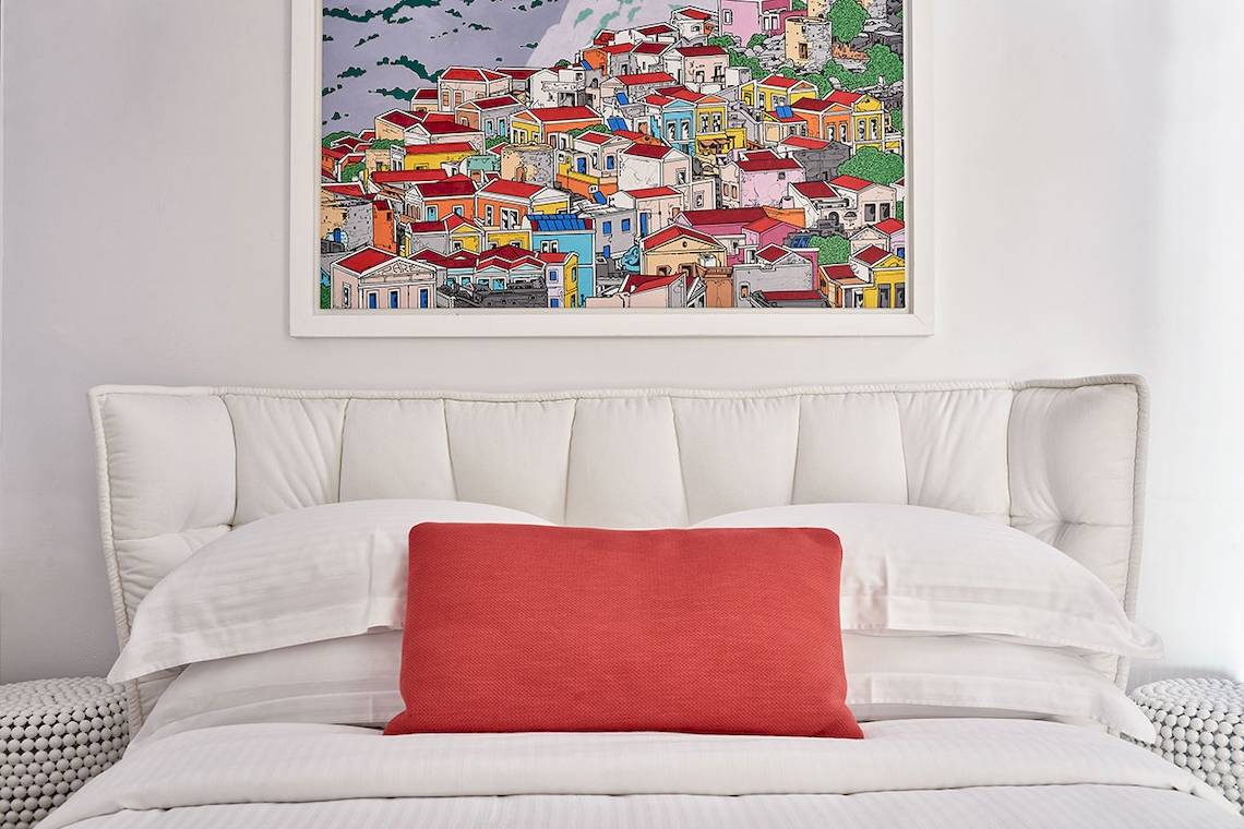 Bett mit weißen Kissen und roter Kopfstütze im Schlafzimmer mit einem Wandbild des Myconian Ambassador Hotels.