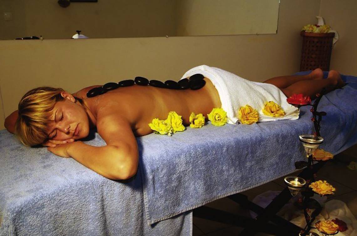 Eine Frau liegt mit geschlossenen Augen auf dem Rücken auf einem Massagetisch mit schwarzen Steinen und gelben Rosen im Hotel.