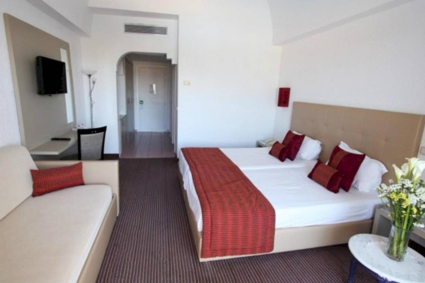 Hotelzimmer im Djerba Holiday Beach mit Doppelbett, Sofa, Fernseher und Tisch mit Blumen.