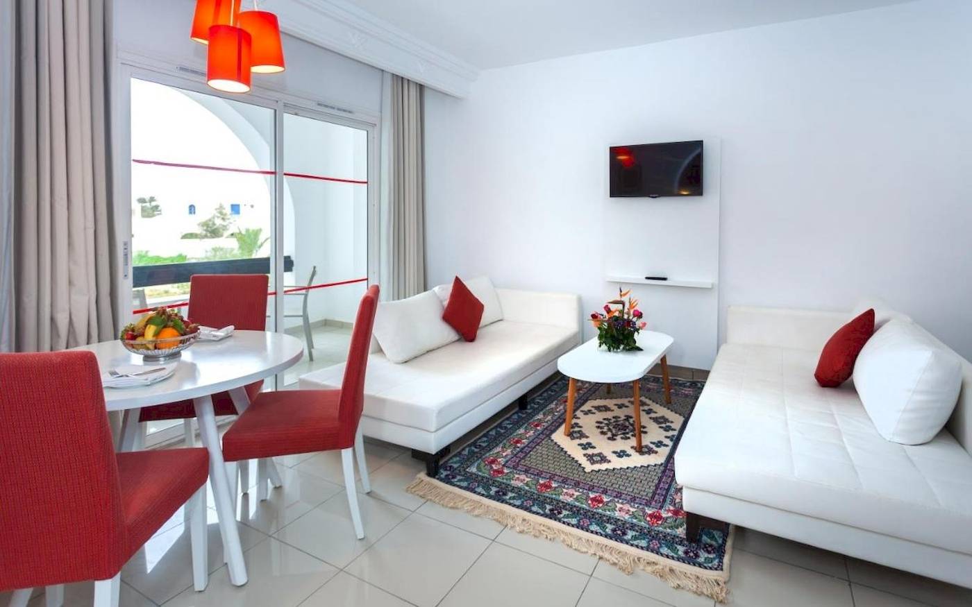 Wohnzimmer im Djerba Holiday Beach Hotel mit weißen Möbeln, rotem Stuhl, Tisch und Fernseher an der Wand.