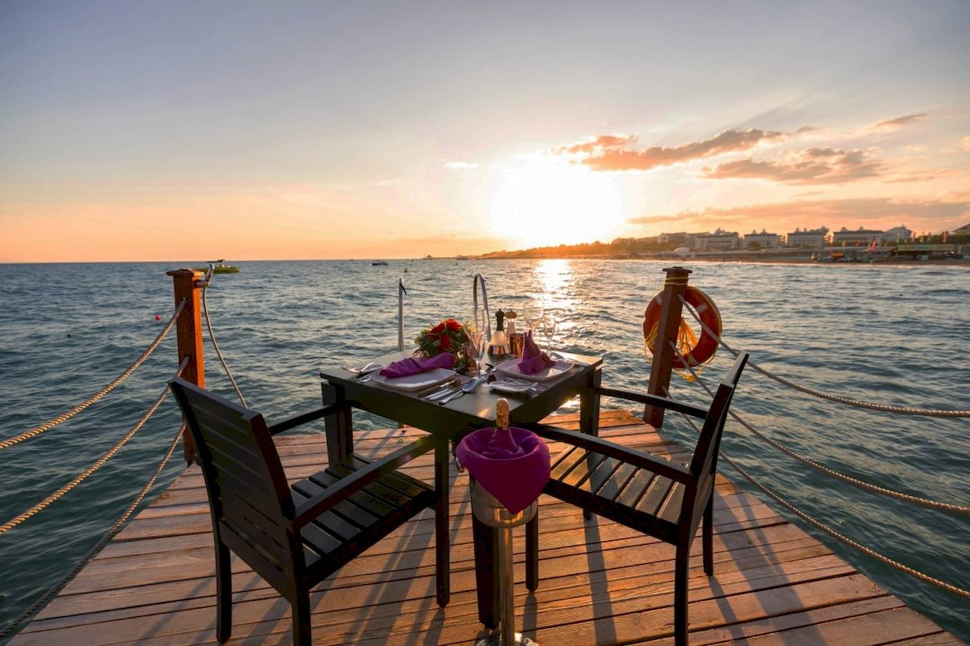 Ein Tisch auf einer Holzplattform am Wasser bei Sonnenuntergang, mit Flaschen und Blumenarrangement.