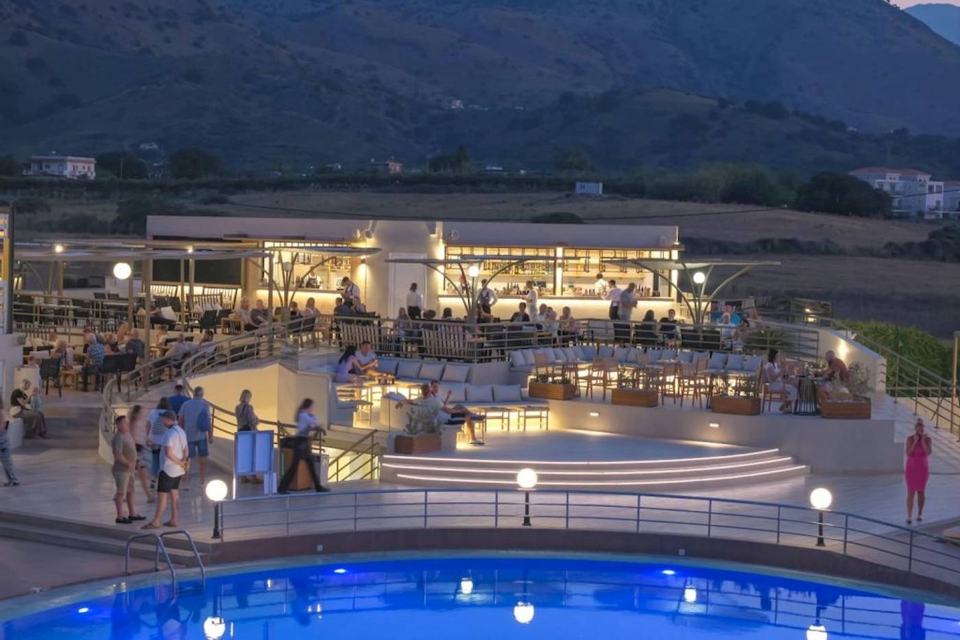 Abend-Pool-Bereich des Georgioupolis Resort Aquapark & SPA mit vielen Gästen und einer Berglandschaft im Hintergrund.