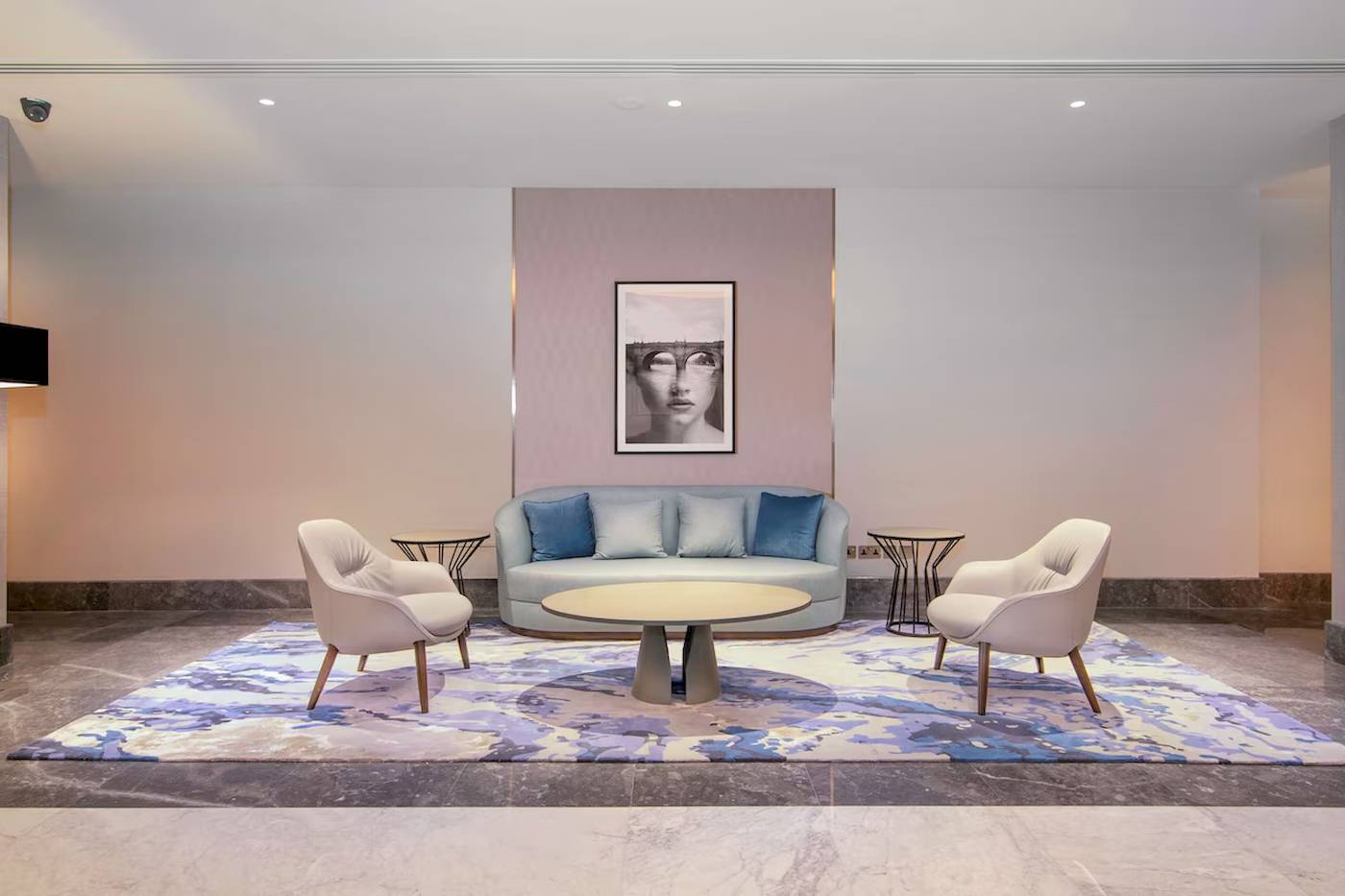 Ein modernes Hotel-Lobby mit einem blauen Sofa, weißen Sesseln und einem zentralen Tisch auf einem großen Teppich.