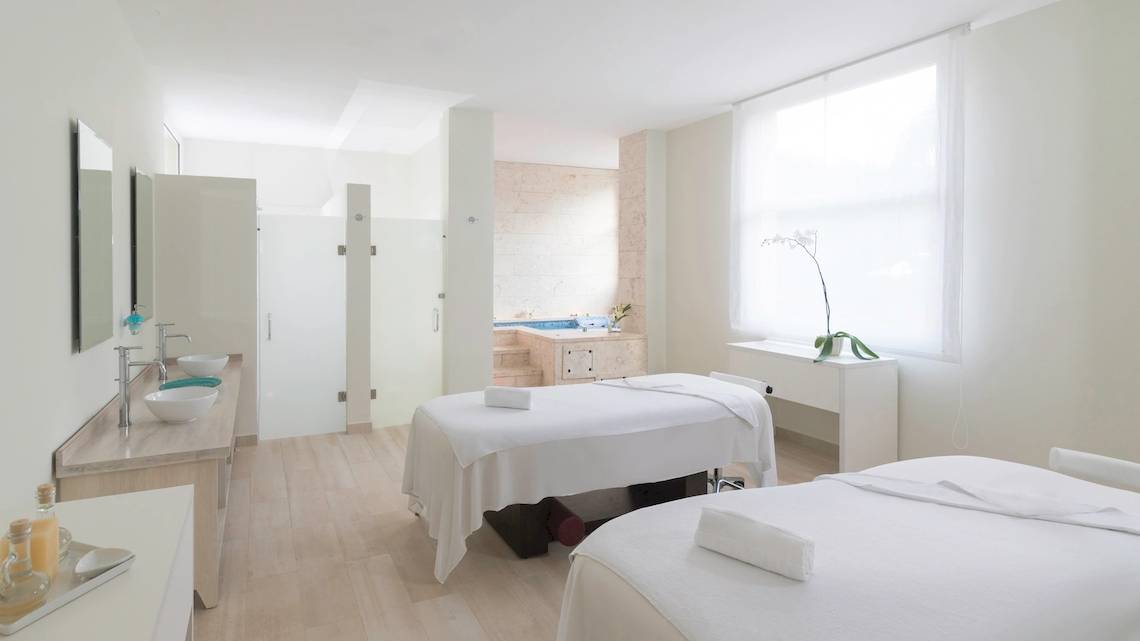 Spa-Behandlungsraum im Hotel JOIA Bávaro by Iberostar mit zwei weißen Behandlungsliegen und einem Whirlpool im Hintergrund.