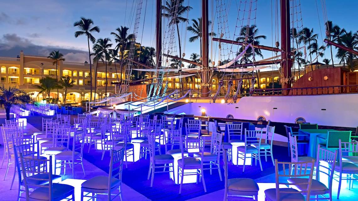 Abendliche Feier auf der Terrasse des JOIA Bávaro by Iberostar Hotel mit blau beleuchteten Stühlen und einem Schiff im Hintergrund.