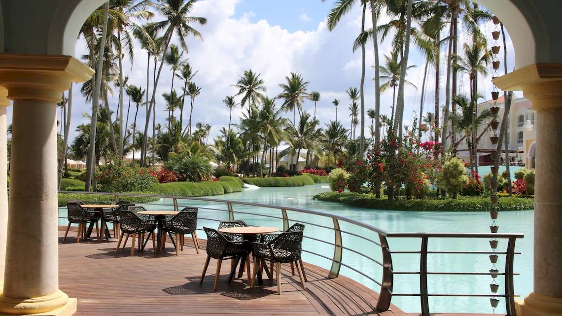 Außenterrasse des JOIA Bávaro by Iberostar mit Tischen und Stühlen und Blick auf einen Wasserweg und Palmen.