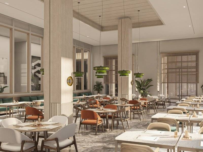 Innenansicht des Restaurants im Jaz Elite Amara Hotel mit runden Tischen und Stühlen, grünen Pflanzen und Kronleuchtern.