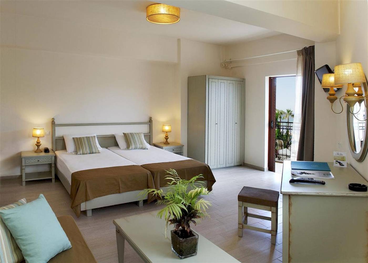 Doppelzimmer im Almyrida Beach Hotel mit zwei Betten, Nachttischen, einer Kommode, einer Lampe und einem offenen Balkon.