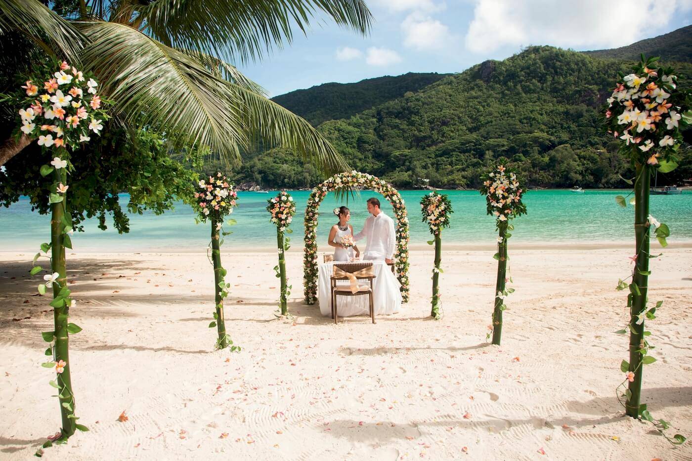 Ein Bräutigam und eine Braut stehen unter einem blumengeschmückten Bogen auf dem Strand beim Constance Ephelia Resort.