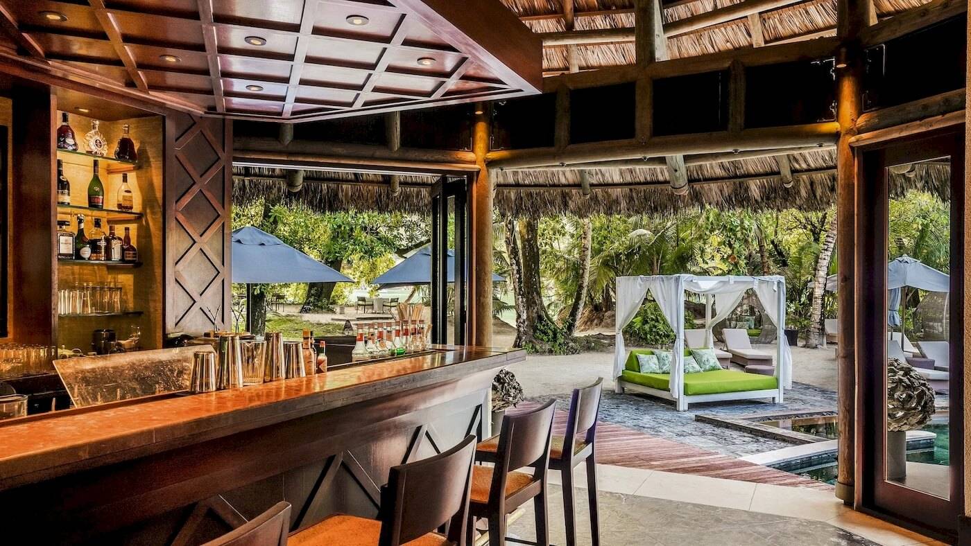 Innenansicht der Bar und Loungebereich des Constance Ephelia Resorts mit grüner Bettlandschaft und Sonnenschirmen.