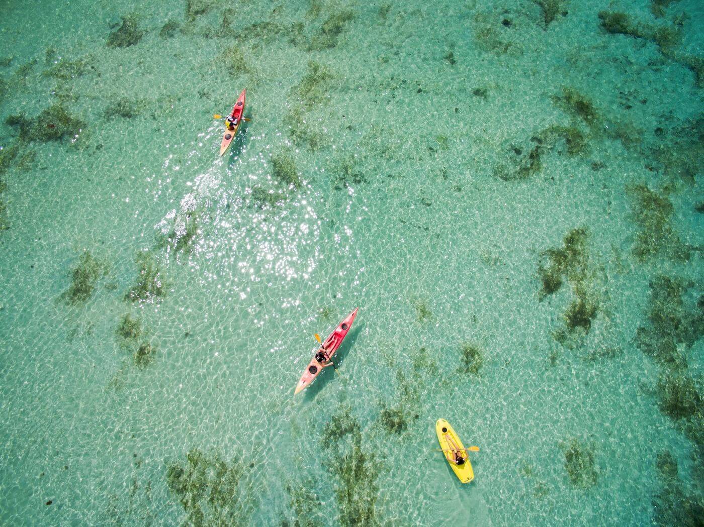 Luftaufnahme von drei Kajakfahrern im kristallklaren Wasser des Constance Ephelia Resorts.