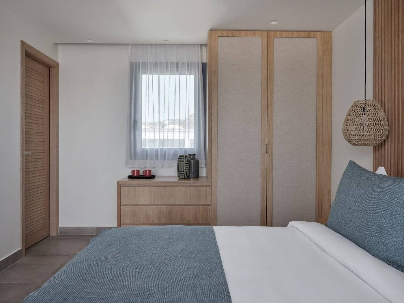 Inneneinrichtung eines Schlafzimmers im Atlantica Beach Resort Kos mit Doppelbett, Nachttisch, Kleiderschrank und Fenster mit Vorhängen.