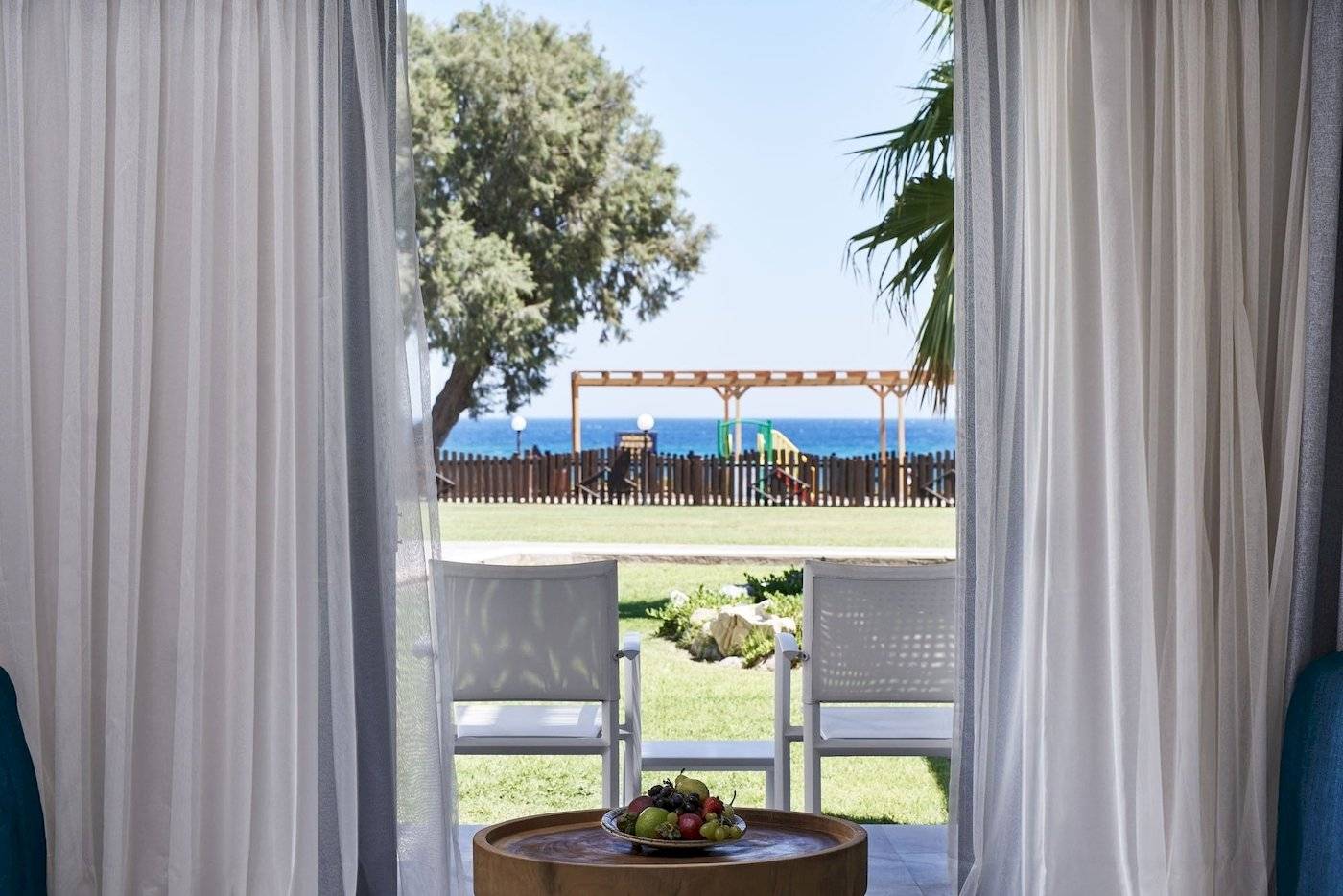 Innenansicht des Atlantica Beach Resort Kos mit Blick auf die Terrasse, auf der zwei weiße Stühle und ein runder Tisch mit Obst stehen.