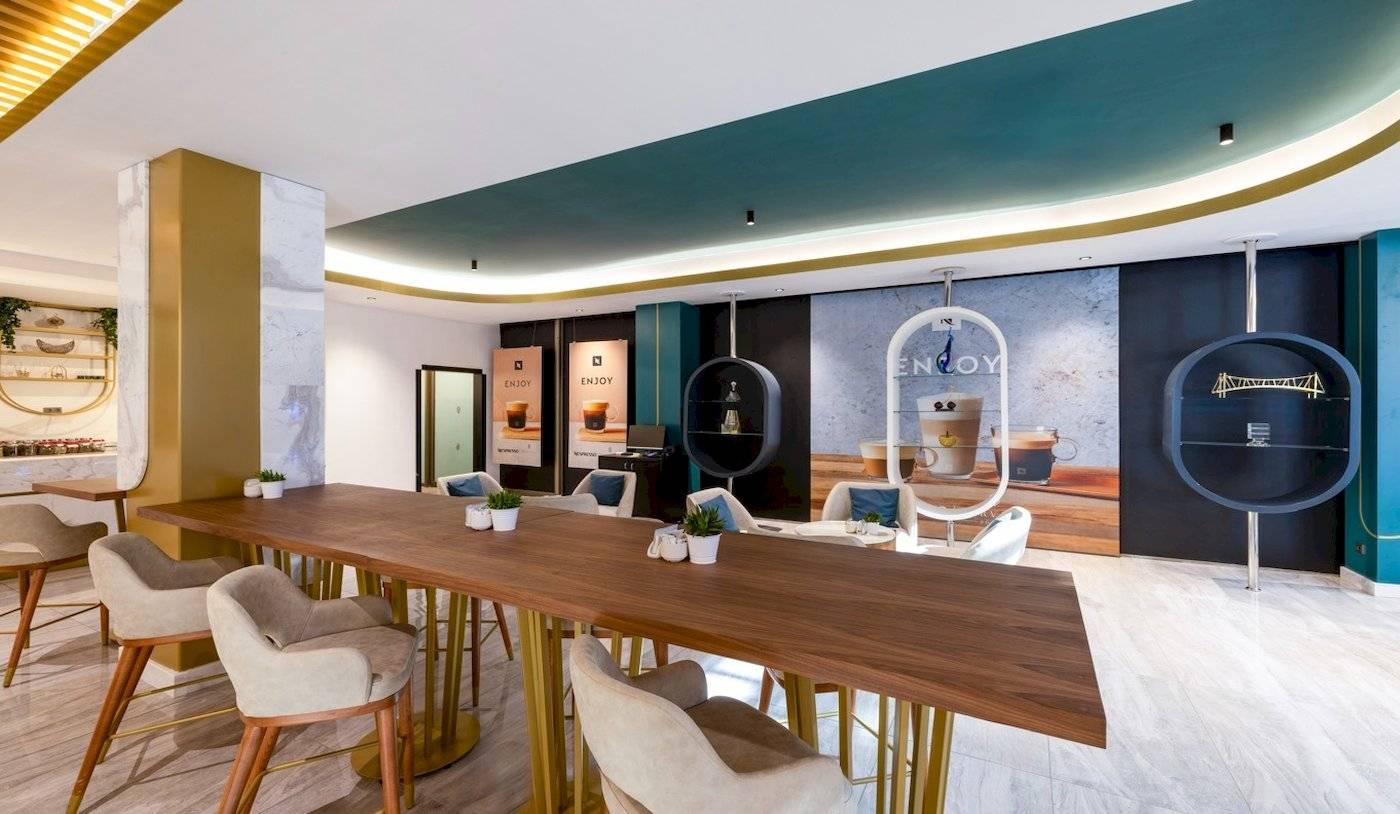 Innenbereich des Nirvana Cosmopolitan Hotels mit einem langen Holztisch, Sesseln, Pflanzen und einer Wand mit einer Wanddekoration.