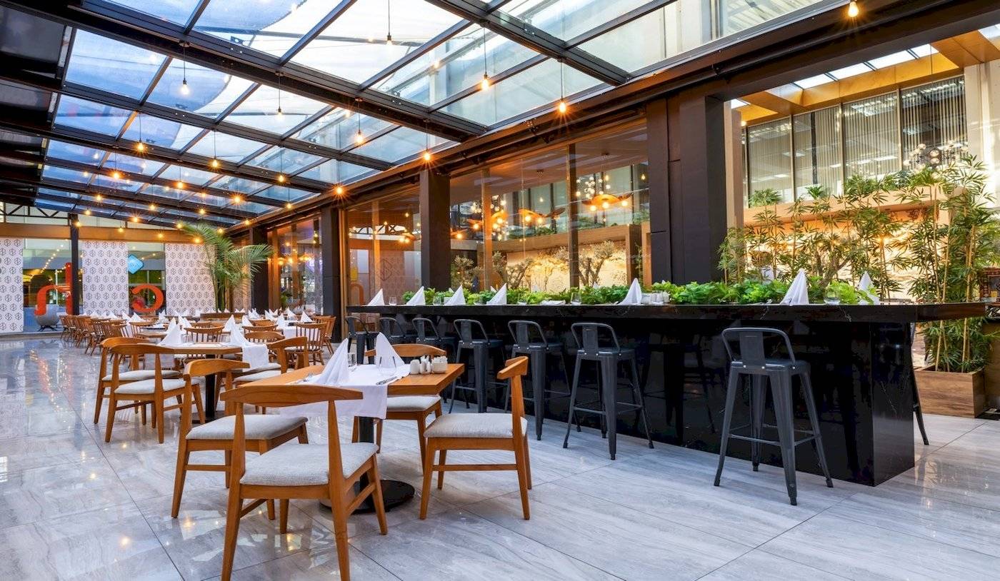 Innenbereich des Nirvana Cosmopolitan Hotels mit Glasdach, Tischen, Stühlen und Pflanzen.