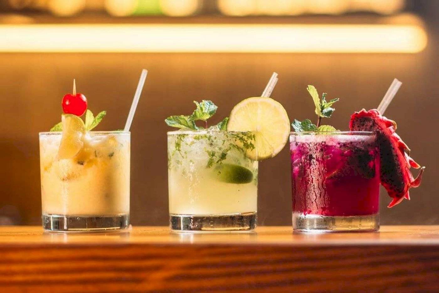 Drei Cocktails auf einem Holztisch, jeder mit verschiedenen Dekorationen wie Früchten und Kräutern.