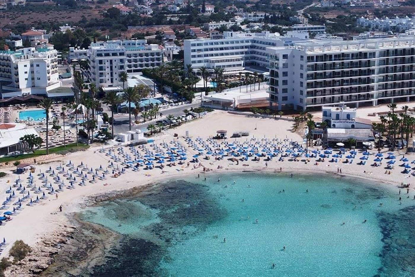 Luftaufnahme des Anonymous Beach Hotels mit vielen Gästen am Strand und blauen Sonnenschirmen.