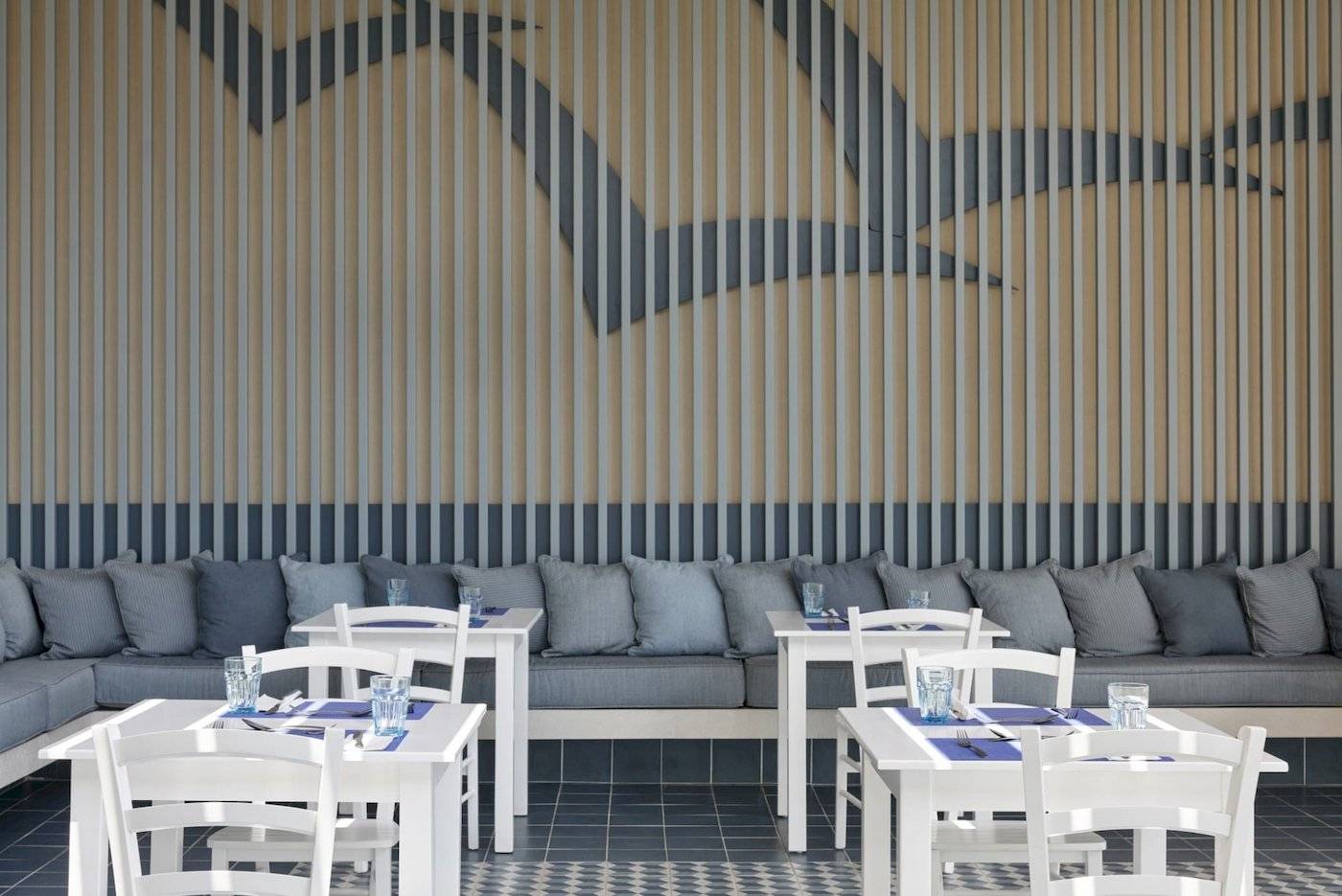 Restaurantbereich im Atlantica Kalliston Resort mit weißen Tischen und Stühlen, grauen Kissen und blauen Wanddekorationen.