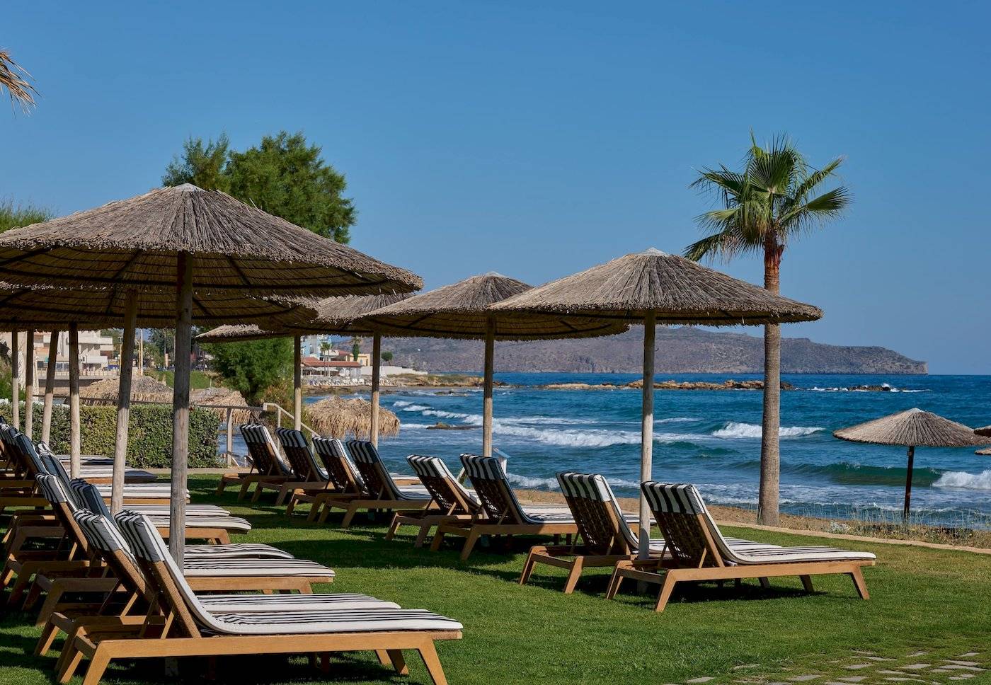 Strandliegen unter Strohhüten am Atlantica Kalliston Resort mit Blick auf das Meer.