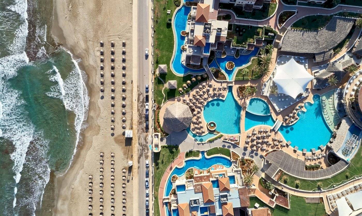 Luftaufnahme des Außenbereichs des Atlantica Kalliston Resorts mit mehreren Swimmingpools und Sonnenschirmen.