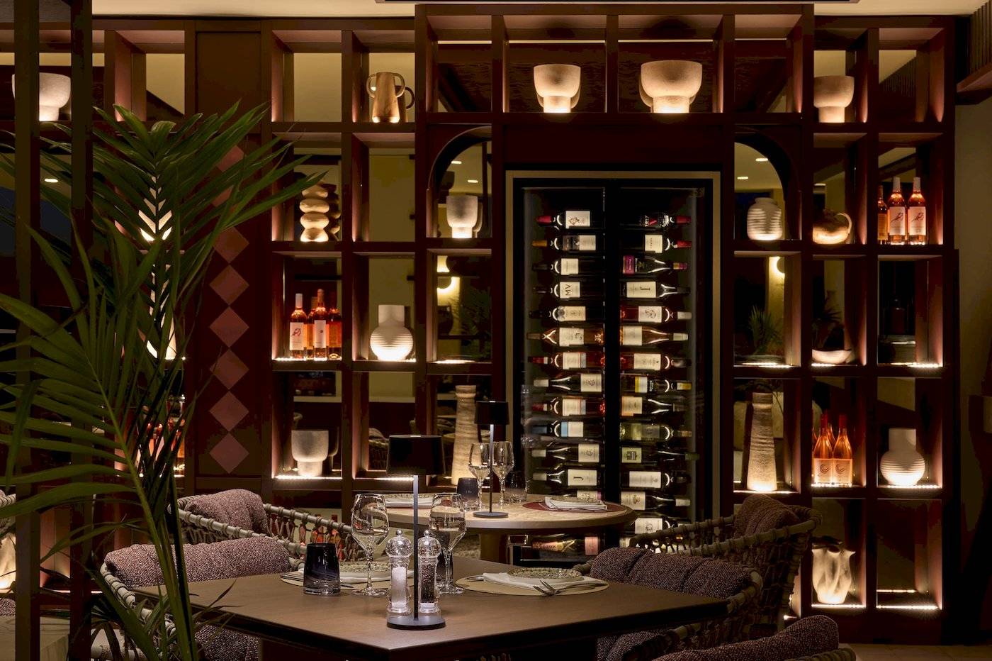 Das Interieur des Atlantica Kalliston Resorts zeigt einen eleganten Essbereich mit braunen Möbeln, Holzregalen und beleuchteten Weinflaschen.