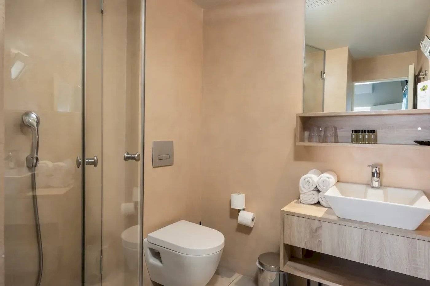 Badezimmer im Corissia Beach Hotel mit Glasdusche, Toilette, Waschbecken und Spiegel.