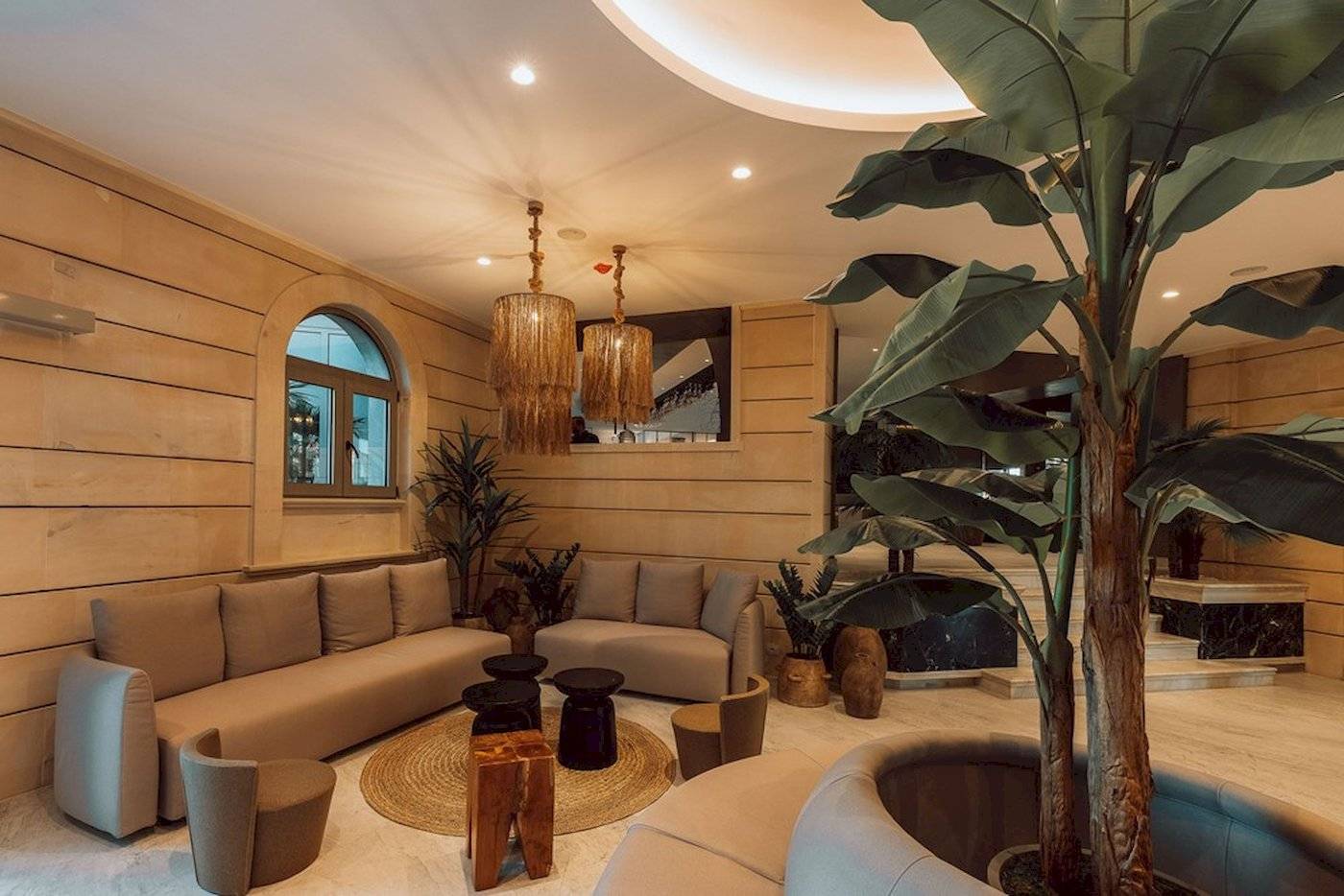 Loungebereich im Eliros Mare Hotel mit beigefarbenen Sofas, Holztischen, Pflanzen und Kronleuchtern.