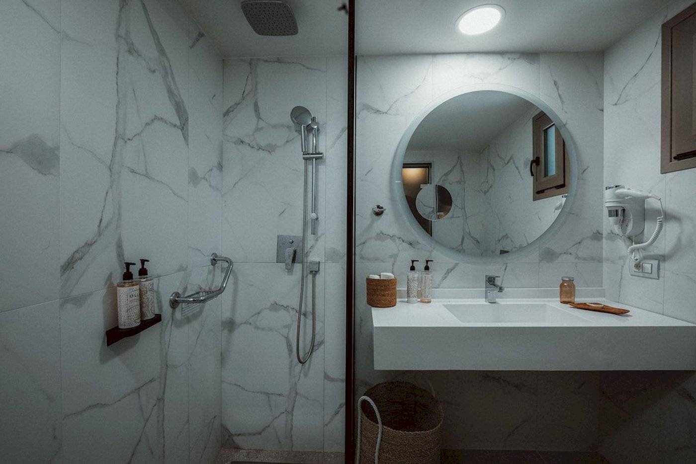 Ein Badezimmer im Eliros Mare Hotel mit einer weißen Marmorwand, einer Glasdusche, einem Spiegel und einem Waschbecken.