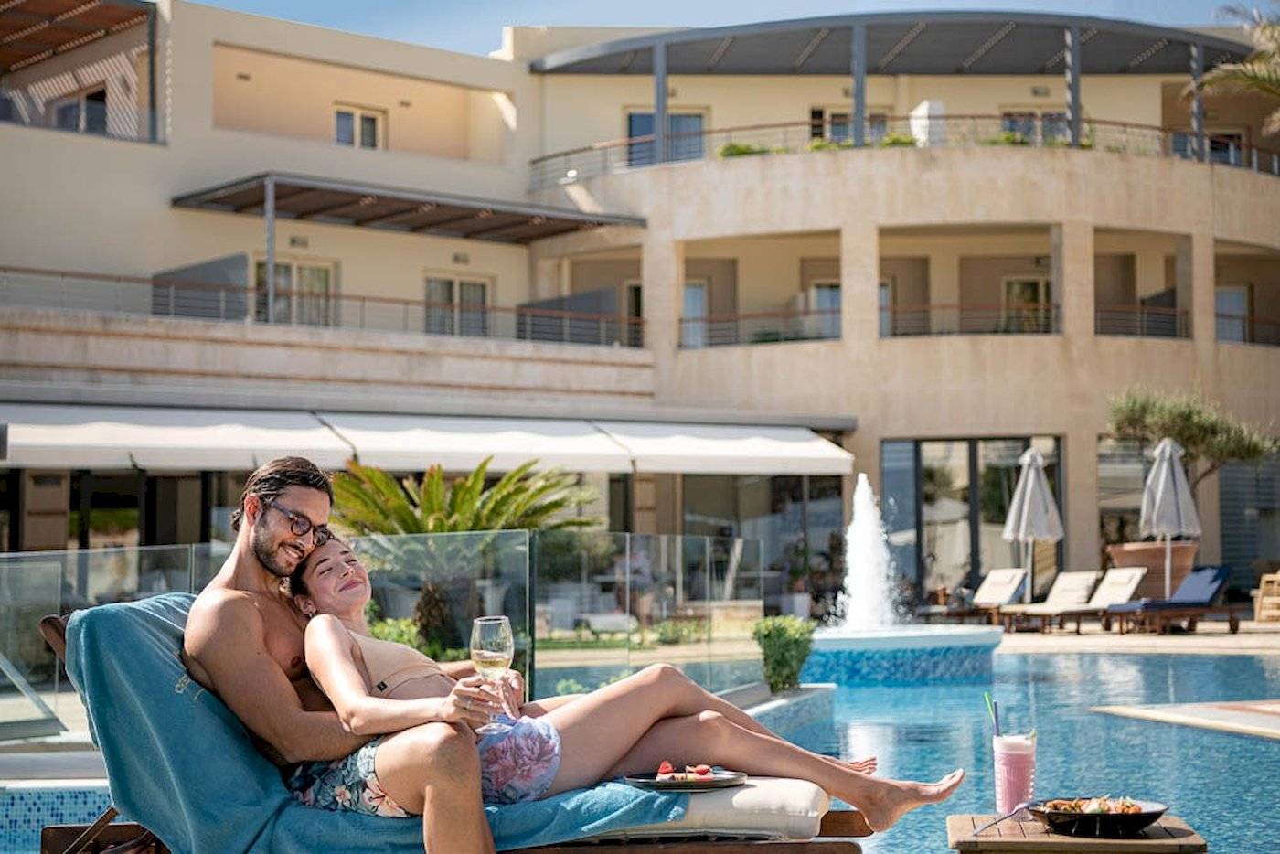Paar am Pool im Cretan Dream Resort & Spa, beide lächelnd, mit einem Glas Wein und Essen vor ihnen.