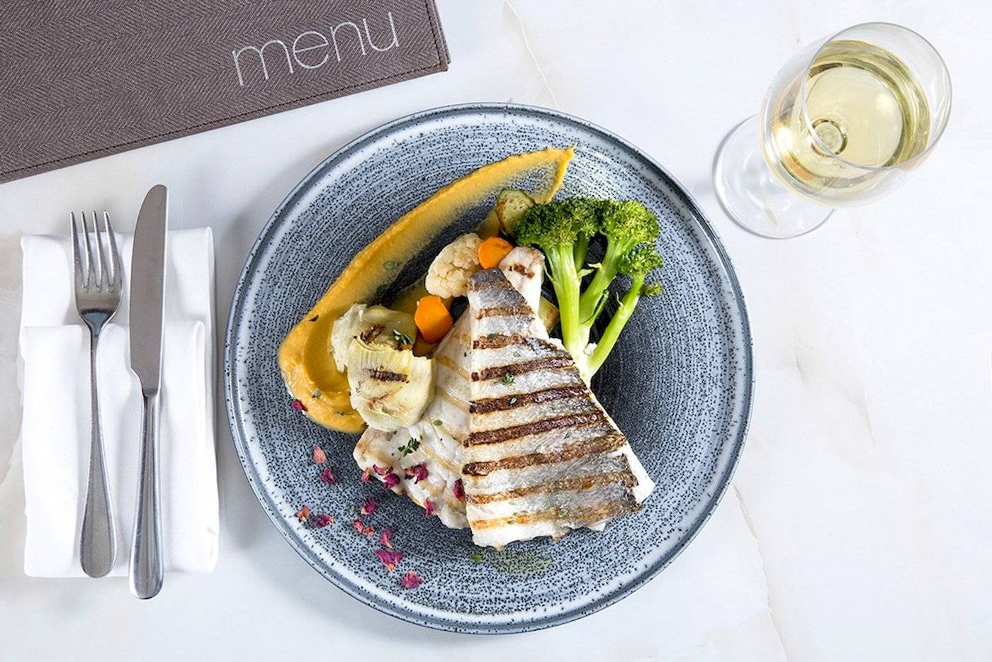 Gegrillter Fisch auf Teller mit Gemüse und Weingläsern auf weißem Tisch