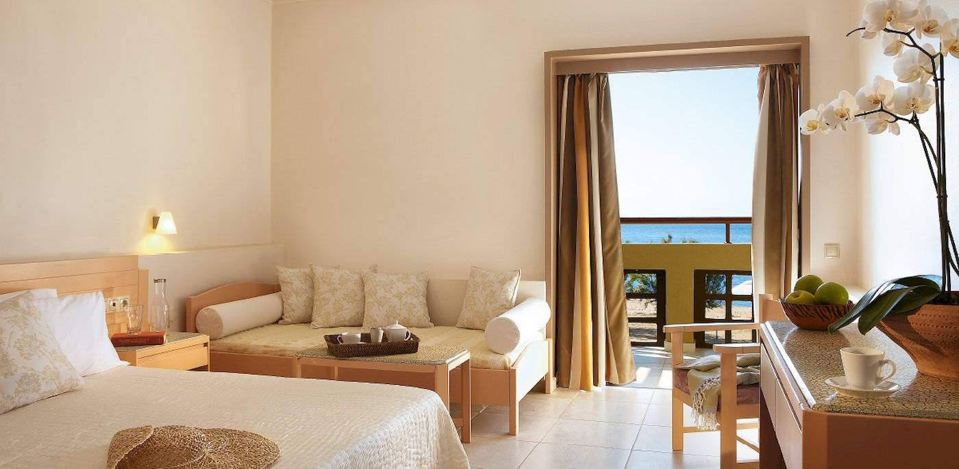 Grecotel Meli Palace Zimmer mit Sofa und Meerblick, das Sofa ist mit Kissen ausgestattet und es gibt einen Tisch mit einer Tasse, einen Nachttisch und eine Vase mit weißen Orchideen.
