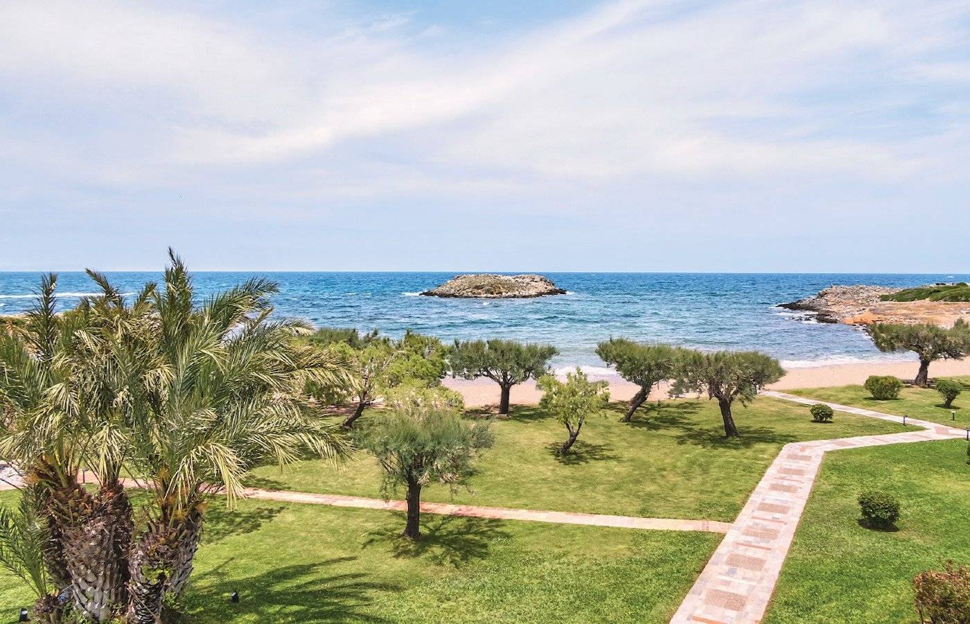 Gesamtansicht des Grecotel Meli Palace mit Blick auf einen Strand und einen kleinen Felsen im Meer.