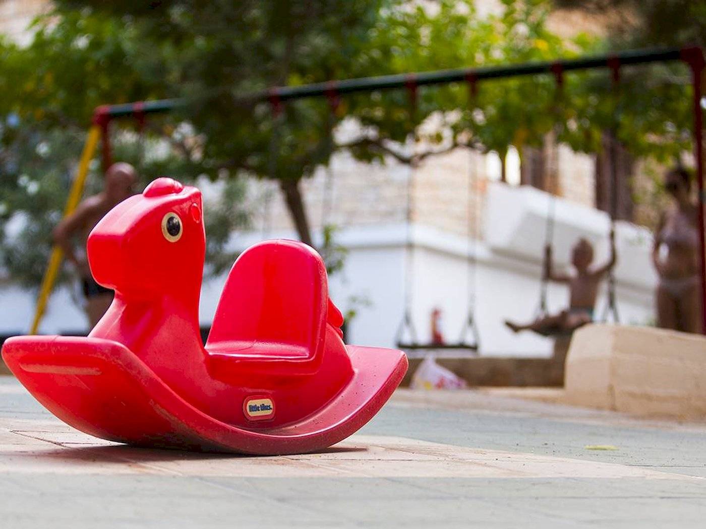 Ein rotes Schaukelpferd im Freien, im Hintergrund sind Kinder auf Schaukeln zu sehen.