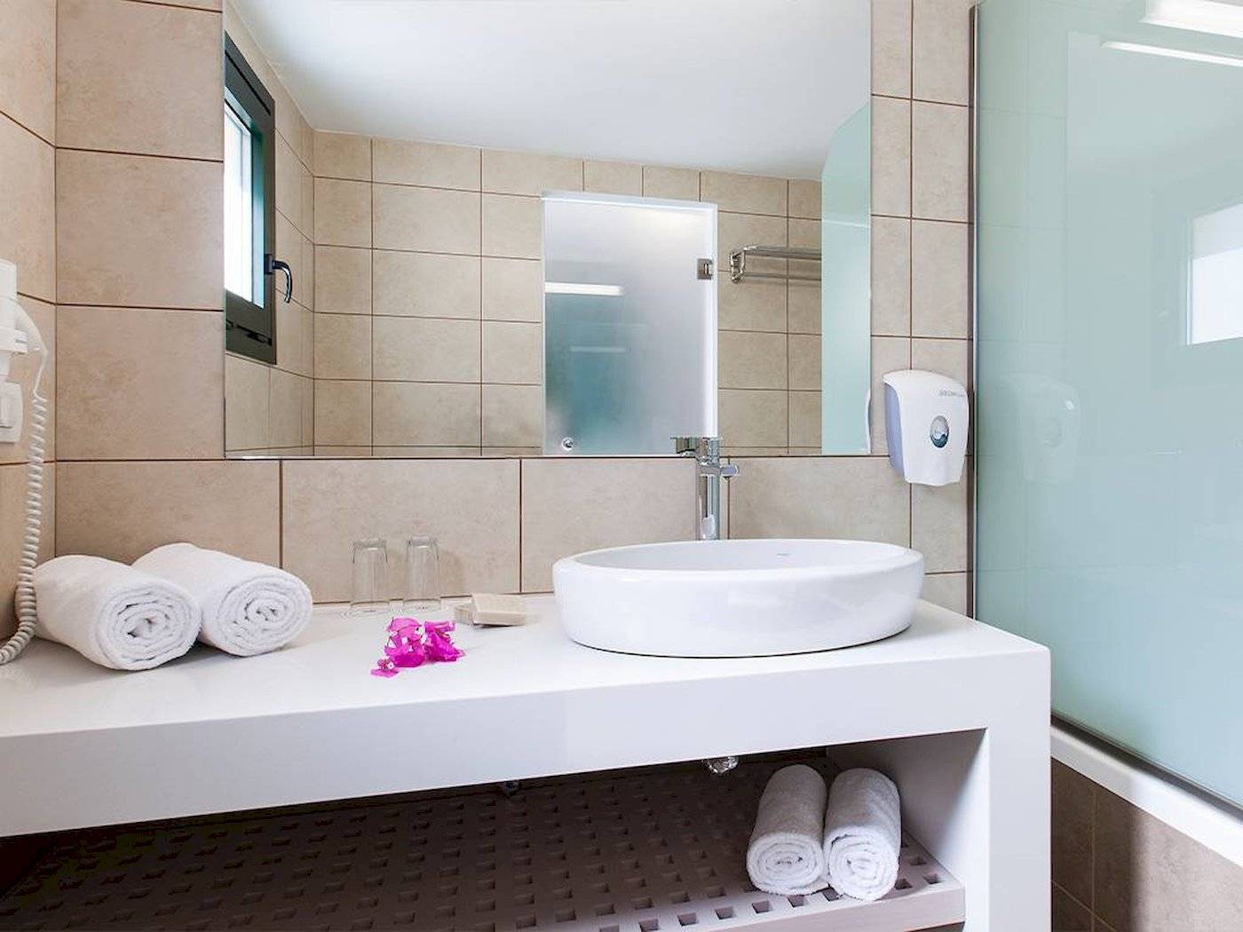 Badezimmer im Chrissi Amoudia Hotel mit Waschbecken, Spiegel und Handtüchern.