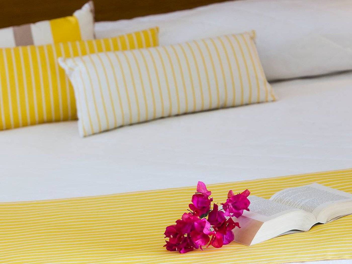 Foto von einem Bett im Chrissi Amoudia Hotel mit gelben Kissen und einer offenen Buch mit Blumen darauf.