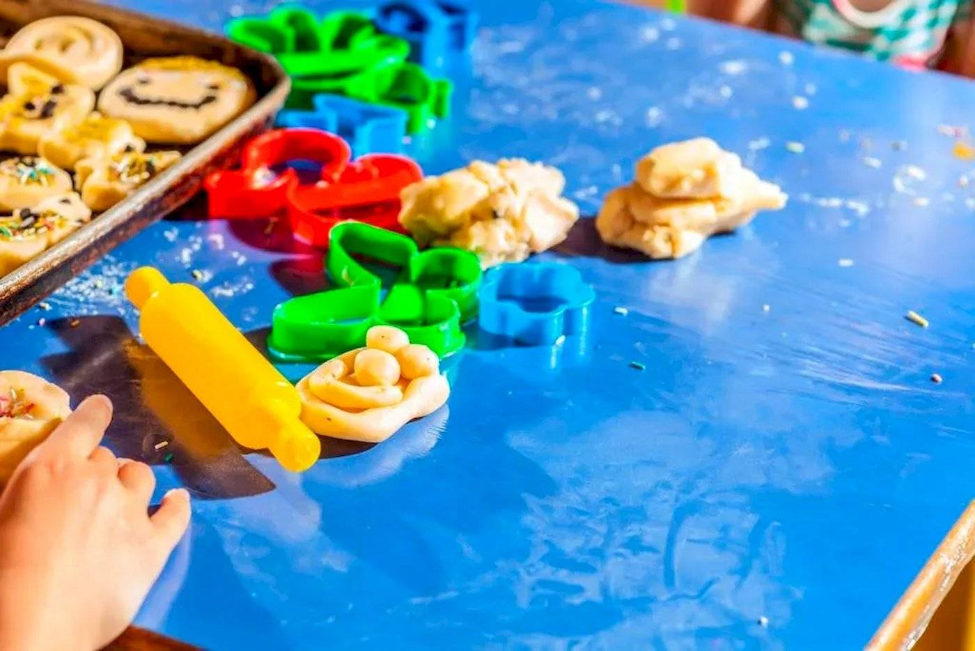Kinder backen Kekse mit Ausstechern und einer Walze auf einem blauen Tisch.