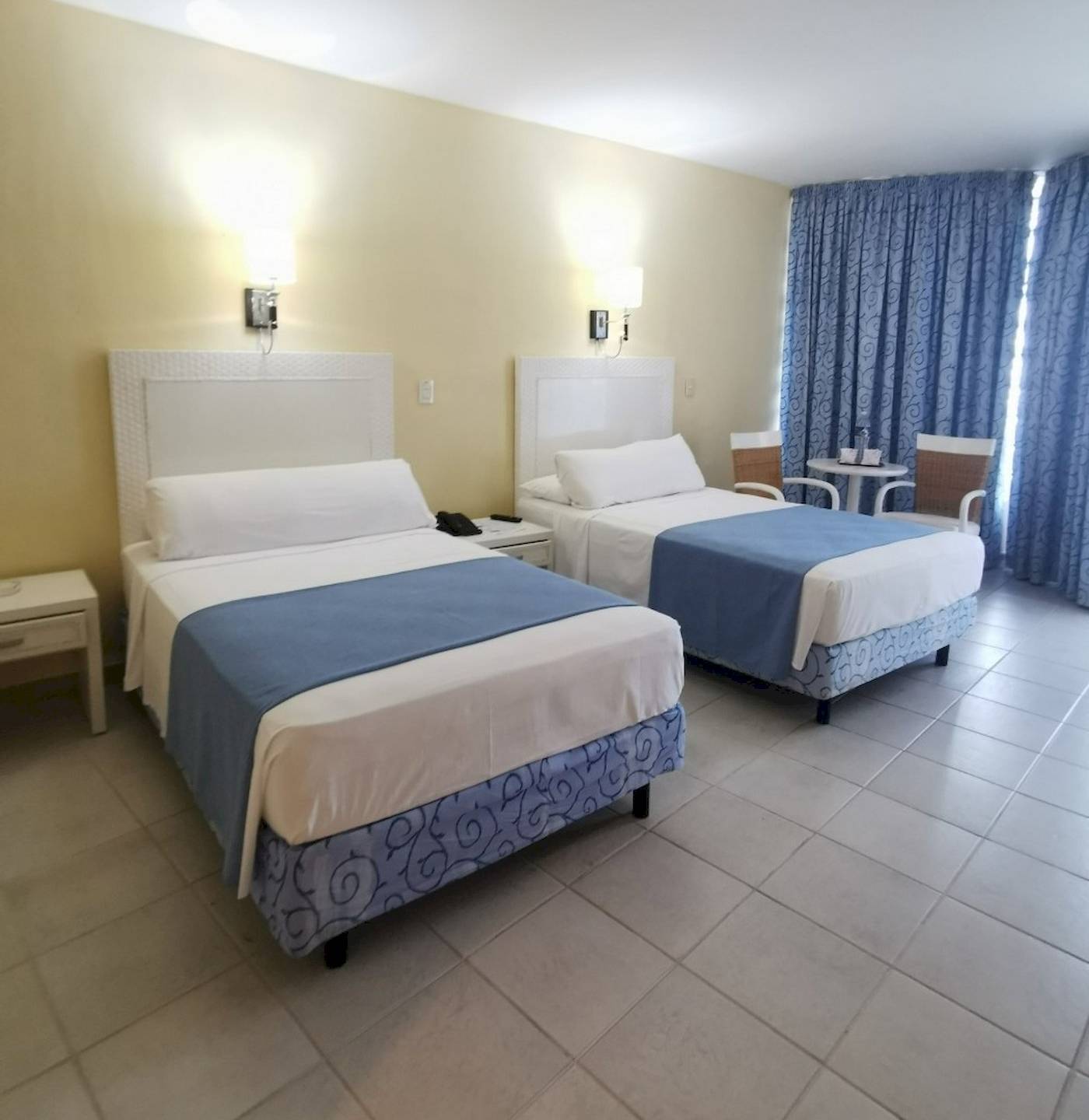 Zwei Doppelbetten im Zimmer des Muthu Playa Varadero Hotels, jedes mit weißen und blauen Laken, Nachttischen und Lampen an der Wand.