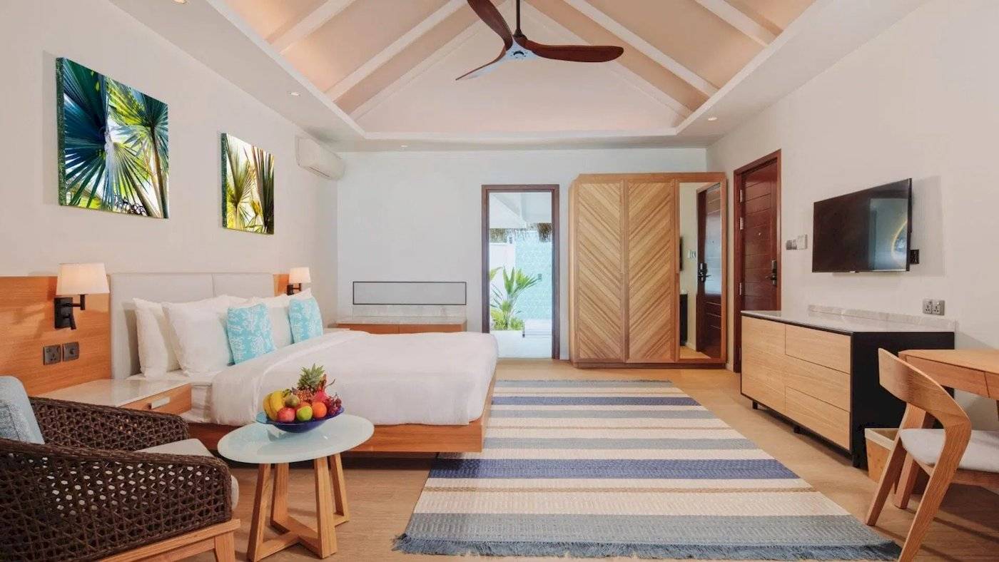 Innenansicht der Nova Maldives Suite mit großem Bett, Früchtetablett, Holzmöbeln und Fernseher.