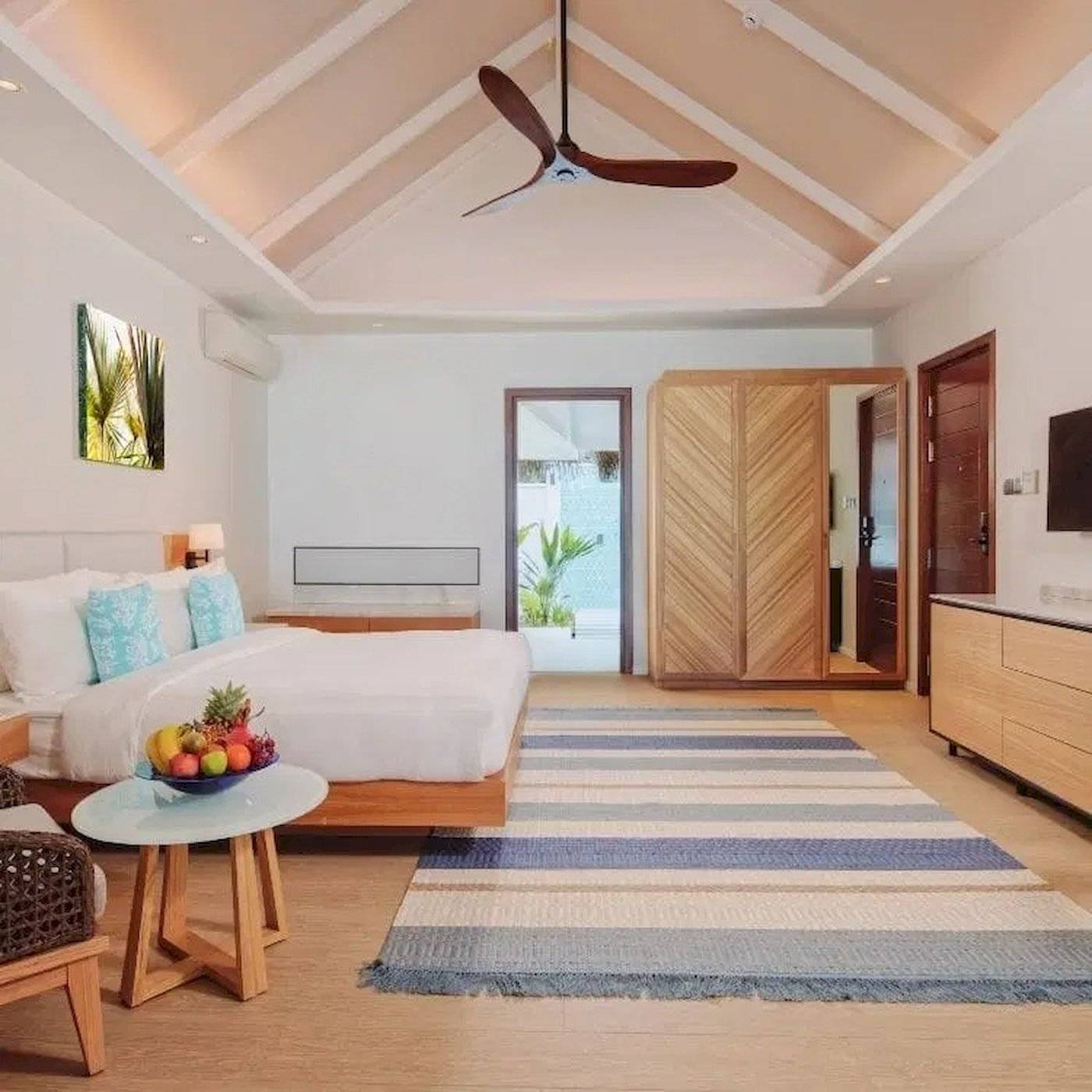 Schlafzimmer im Nova Maldives Hotel mit weißem Bett, Holzmöbeln und blau-weiß gestreiftem Teppich.