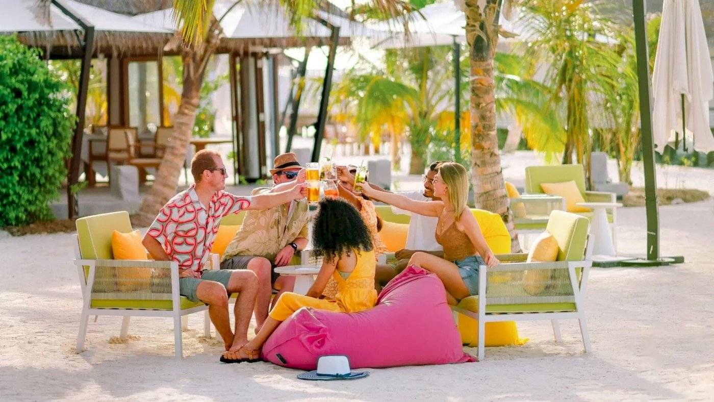 Gäste des Nova Maldives Hotels sitzen am Strand und chören mit Getränken in der Hand.