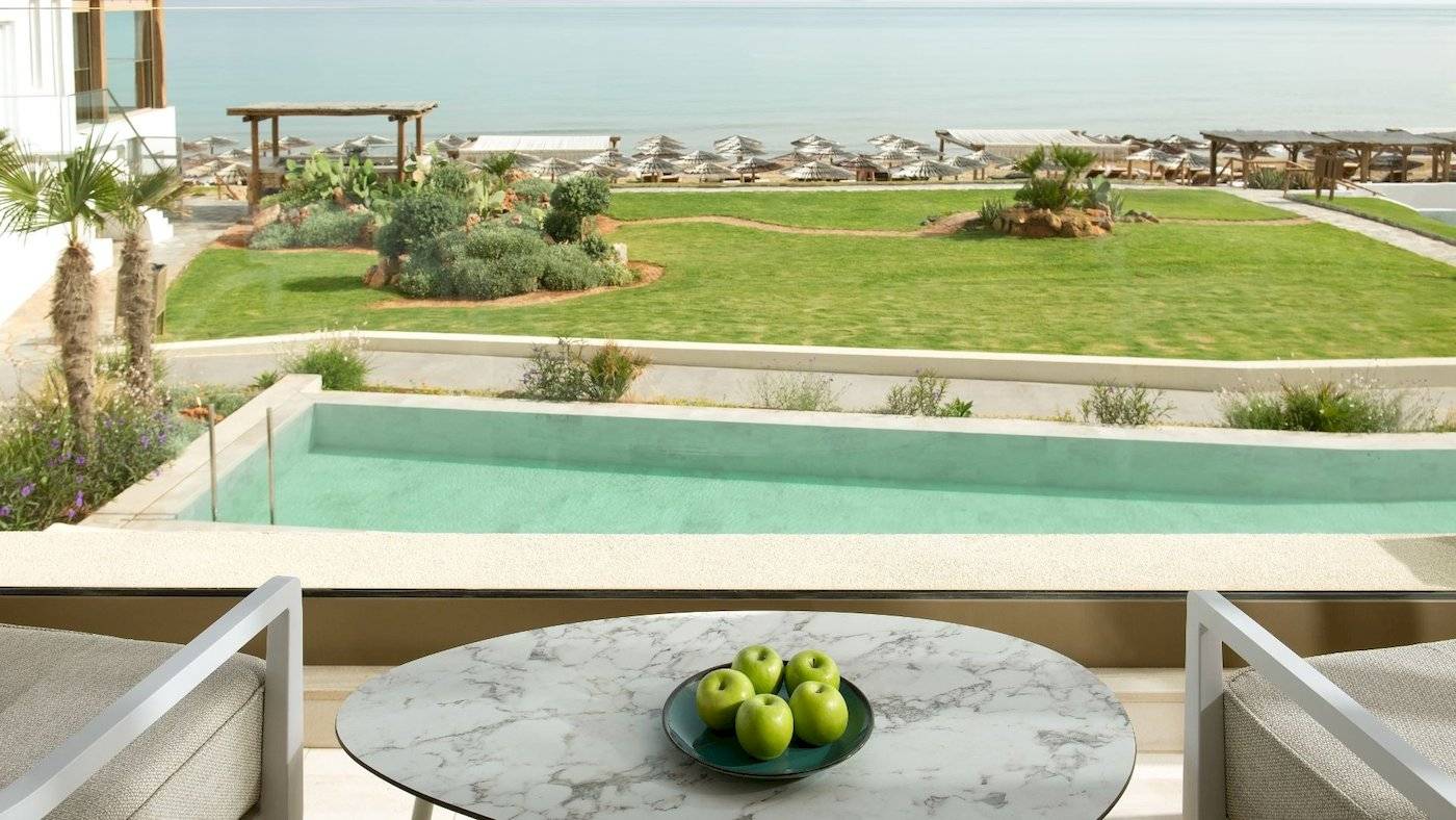 Blick vom Balkon des Mitsis Selection Rinela Hotels auf eine Terrasse mit Tisch, Äpfeln und Blick auf den Pool und das Meer.