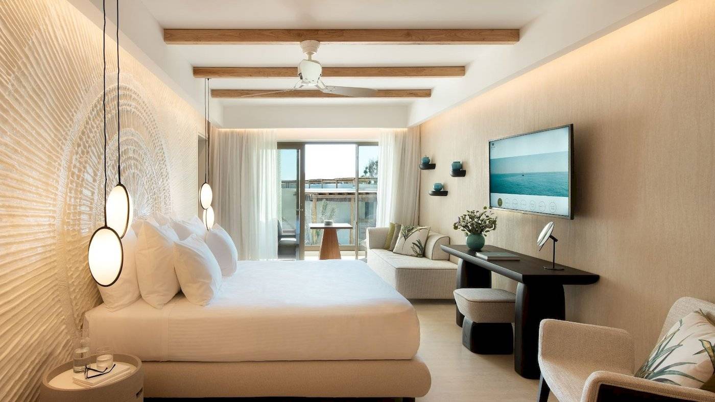 Innenansicht eines Hotelzimmers im Mitsis Selection Rinela mit Bett, Couch, Schreibtisch und Fernseher.