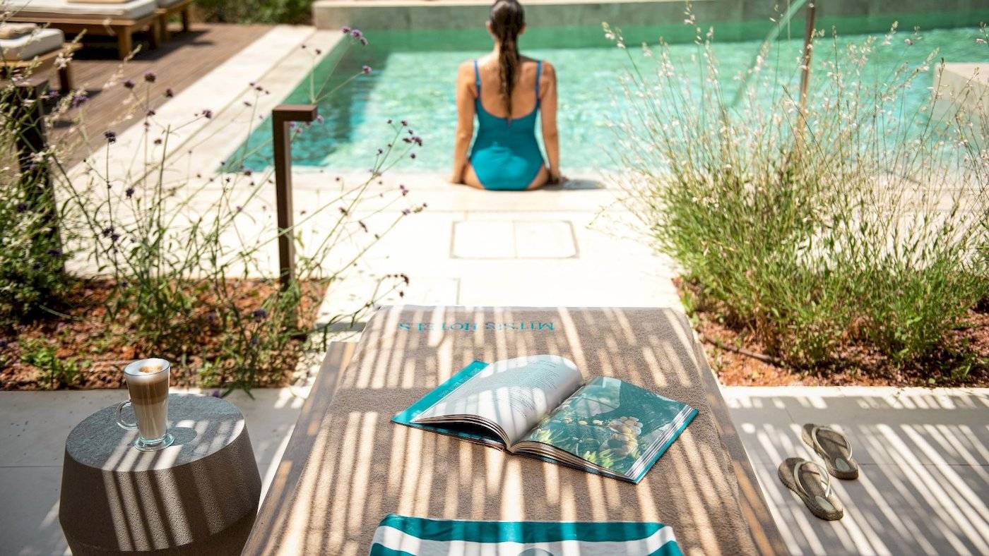 Eine Frau in einem blauen Badeanzug sitzt an einem Pool am Rand und liest ein Buch.