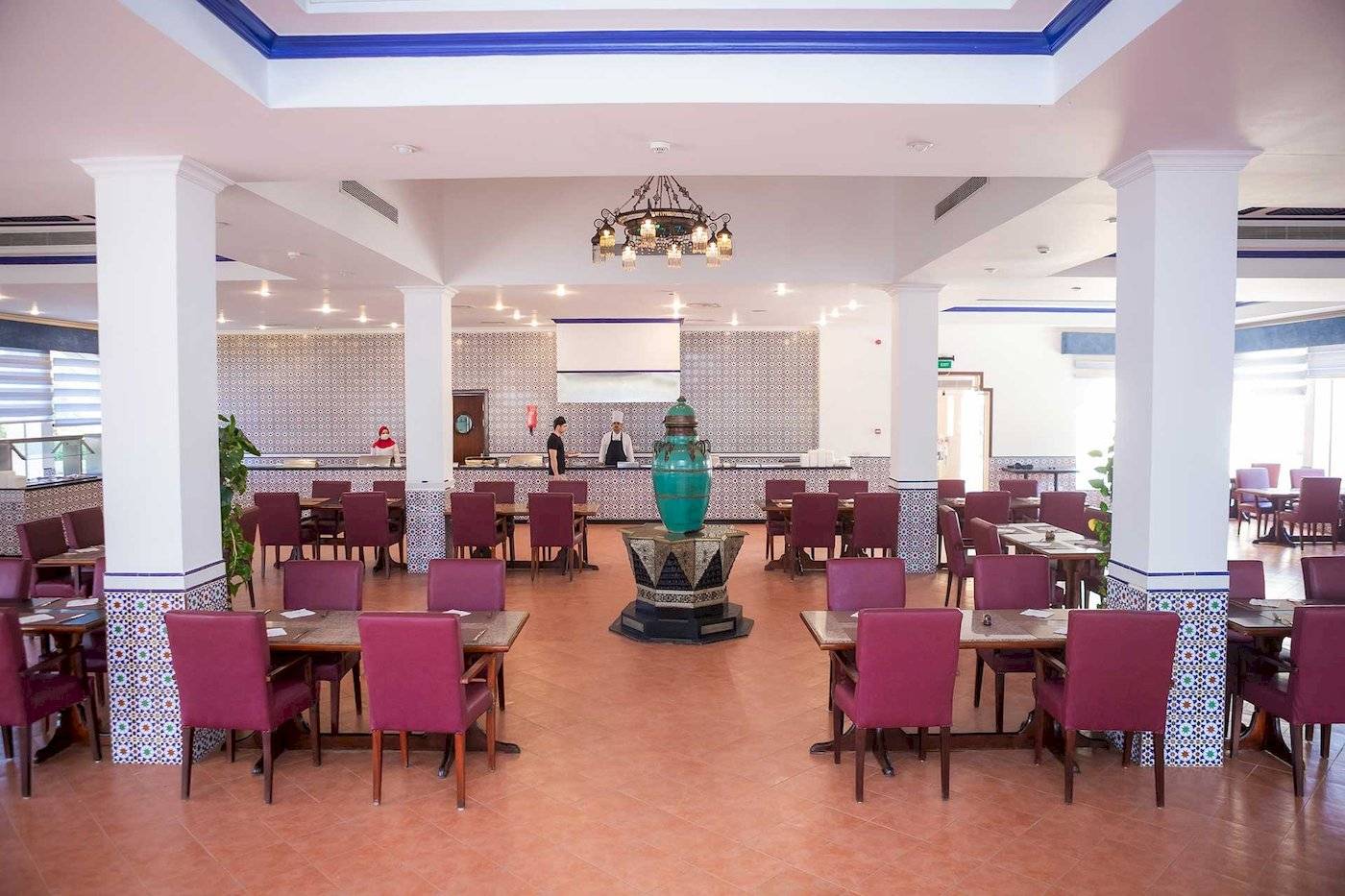 Innenbereich des Stella Gardens Resort & Spa Makadi Bay mit geometrischen Wandverkleidungen, runden Tischen, roten Stühlen, grünem Brunnen und hängender Lampe.